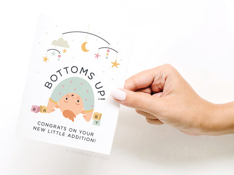 A hand holds the Onderkast Studio Bottoms Up Baby Greeting Card, featuring an illustrated baby doing a handstand among baby blocks and a mobile. The card reads, BOTTOMS UP! Congrats on your new little addition!.