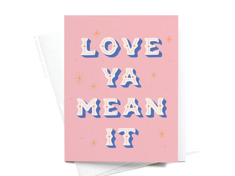 The Love Ya Mean It Greeting Card by Onderkast Studio features retro-style white and blue text on pink, accented with small stars. A partially visible envelope enhances this charming design’s premium look.