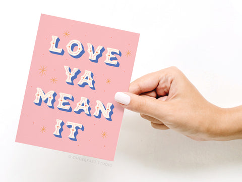 A hand with light pink nail polish holds Onderkast Studio’s Love Ya Mean It Greeting Card—a pink card featuring bold white and blue “LOVE YA MEAN IT” text, accented with small star illustrations.