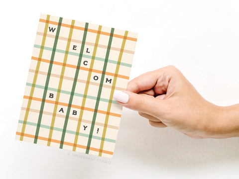 A hand with light pink nail polish holds the Welcome Baby Gingham Greeting Card by Onderkast Studio, featuring a green, orange, and yellow gingham pattern and “WELCOME BABY!” in bold black letters arranged vertically and horizontally.