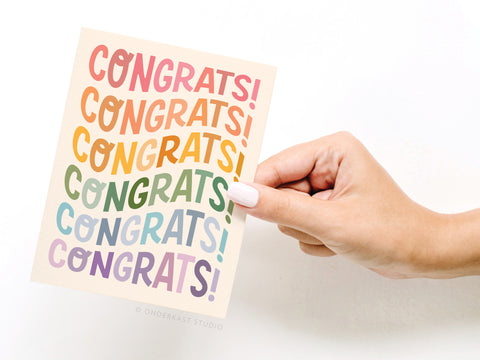 A hand with light pink nail polish holds the Onderkast Studio Congrats Wonky Type Greeting Card, featuring playful fonts and vibrant CONGRATS! text on a white background for a premium look.