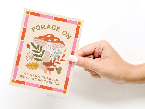 A hand holds the Forage On Mushroom Greeting Card by Onderkast Studio, featuring colorful mushrooms, leaves, and flowers with FORAGE ON and WE GROW THROUGH WHAT WE GO THROUGH in bold within a pink-orange border.