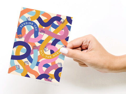 A hand holds Onderkast Studios Sending You All the Hugs Greeting Card, decorated with intertwined pink, blue, yellow, and purple arms and the message “Sending You All the Hugs”—a heartfelt thinking of you card.