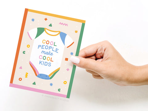 A hand with white nail polish holds the Onderkast Studio Cool People Make Cool Kids Greeting Card, featuring a playful onesie illustration and multicolored text—an ideal greeting card for new parents.