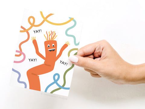 A hand with white nail polish holds the Onderkast Studio Yay Wacky Waving Inflatable Tube Man Greeting Card, featuring a cheerful orange tube man and YAY! written three times, surrounded by colorful squiggly lines.