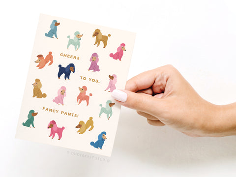 A hand holds the Onderkast Studio Cheers to You Fancy Pants Poodles Greeting Card, featuring vibrant poodles and playful “Cheers to you, fancy pants!” text for a premium touch.
