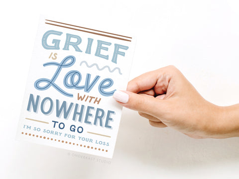 A hand with light pink nail polish holds the Onderkast Studio Grief Is Love With Nowhere to Go Greeting Card—a premium quality sympathy card featuring heartfelt blue and brown fonts, perfect for expressing condolences.