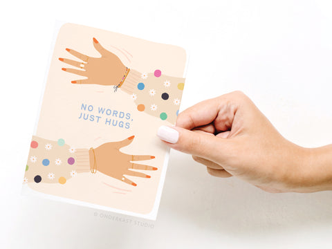 A hand with white nails holds the Onderkast Studio No Words Just Hugs Greeting Card, which features two illustrated arms in polka-dot sleeves and gold bracelets reaching for a hug, with NO WORDS, JUST HUGS in blue letters on the front.