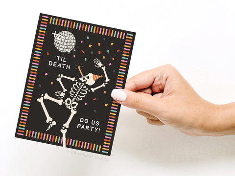 A hand holds Onderkast Studios Til Death Do Us Party Greeting Card, featuring a dancing skeleton in a party hat under a disco ball with confetti and the text TIL DEATH DO US PARTY! on a striped black background.