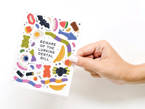 A hand holds Onderkast Studio’s Beware of the Lurking Dental Bill Greeting Card, featuring bright, spooky dental-themed illustrations—candies, teeth, and sweets—for a humorous Halloween twist.
