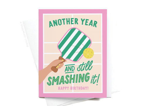 The Another Year and Still Smashing It Pickleball Greeting Card by Onderkast Studio features a pink border, a hand with a green-and-white striped paddle hitting a yellow ball, and cheerful birthday wishes for pickleball fans.