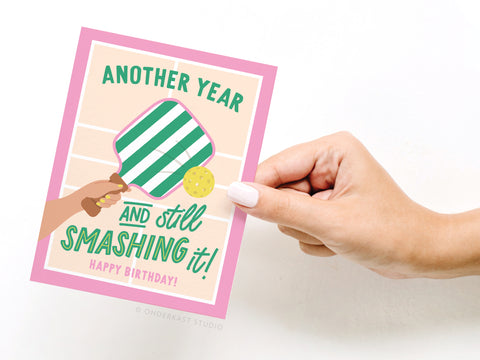 A hand displays Onderkast Studio’s “Another Year and Still Smashing It Pickleball Greeting Card,” featuring a striped paddle and ball with the playful birthday message in bold, premium fonts.
