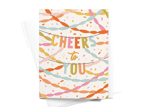 The Cheers to You Party Streamers Greeting Card by Onderkast Studio features bold, colorful streamers and CHEERS TO YOU on a white background—perfect for birthdays. An envelope peeks out from behind the card.