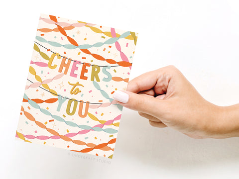 A hand with light pink nail polish holds the Onderkast Studio Cheers to You Party Streamers Greeting Card, which features colorful streamers and bold text saying CHEERS TO YOU.