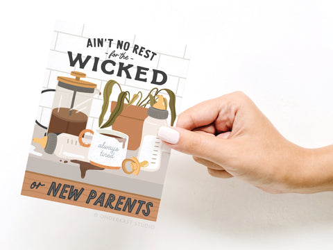 A hand holds the Ain’t No Rest for the Wicked or New Parents Greeting Card by Onderkast Studio, featuring vibrant prints of coffee, a baby bottle, pacifier, spilled coffee, tired plants—an ideal choice for new parents.