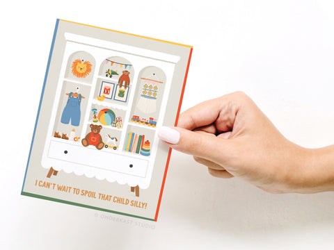 A hand holds the Onderkast Studio “I Can’t Wait To Spoil That Child Silly” greeting card, featuring a colorful cabinet filled with baby essentials and playful items. The caption reads, I cant wait to spoil that child silly!.