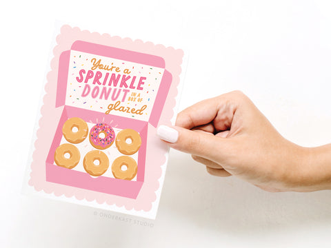 A hand holds the Onderkast Studio Youre a Sprinkle Donut Greeting Card, featuring a pink donut box with five plain donuts and one sprinkled donut, and the message: You’re a SPRINKLE DONUT in a box of glazed.