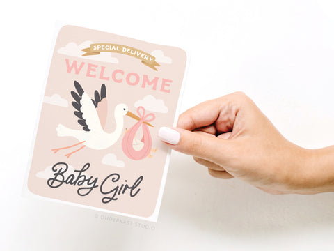 A hand holds the Onderkast Studio Special Delivery Welcome Baby Girl Greeting Card, featuring a flying stork with a pink bundle and the message Special Delivery, Welcome Baby Girl on a soft pink, cloudy background.
