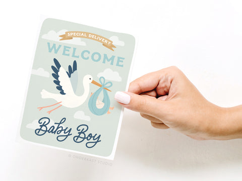 A hand holds the Onderkast Studio Special Delivery Welcome Baby Boy Greeting Card, featuring a vibrant illustrated stork with a blue bundle, cloud background, and the text Special Delivery.