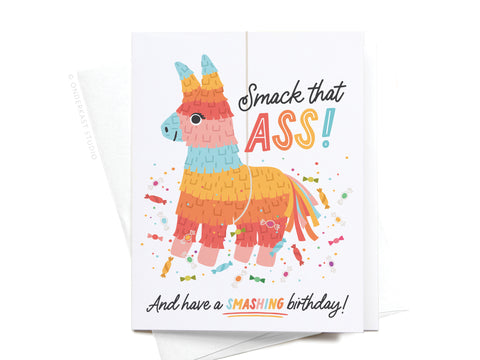 The Smack That Ass Piñata Greeting Card by Onderkast Studio features a donkey-shaped piñata with Smack that ASS! And have a SMASHING birthday! on a white background, surrounded by confetti and candies.