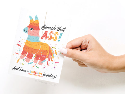 A hand holds the Smack That Ass Piñata Greeting Card by Onderkast Studio, featuring a donkey-shaped piñata and the text Smack that ASS! And have a SMASHING birthday! with confetti and candy illustrations for a festive look.