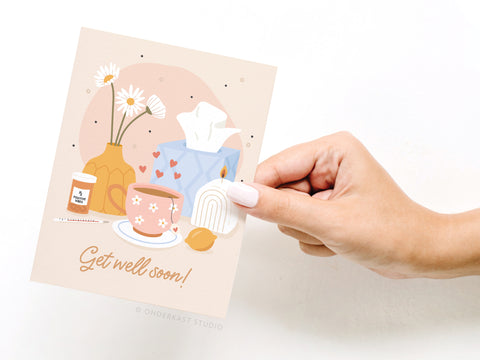 A hand holds the Onderkast Studio Get Well Soon Greeting Card, featuring cozy designs—a vase of flowers, teacup, tissue box, medicine jar, thermometer, lemon, and hearts—on a premium soft pink background.