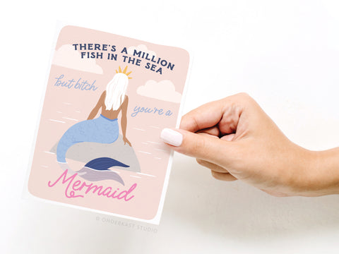 A hand displays the Onderkast Studio There’s a Million Fish in the Sea Mermaid Greeting Card, featuring a crowned mermaid illustration and the phrase, There’s a million fish in the sea, but bitch you’re a mermaid, on soft pink with water and fish.