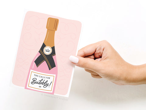 A hand with light pink nail polish holds Onderkast Studio’s “This Calls for Bubbly Greeting Card, featuring a pink champagne bottle on a soft, swirly pink background for a premium look.