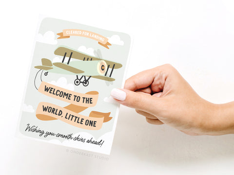 A hand holds the Onderkast Studio Welcome to the World Little One Greeting Card, featuring a vintage airplane and banners with messages like “Cleared for landing” against a cloud-filled sky.