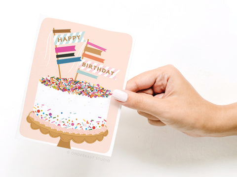 A hand with white nail polish holds the Onderkast Studio Happy Birthday Sprinkle Cake Greeting Card, featuring a birthday cake with colorful flag toppers that spell out Happy Birthday.