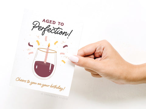 A hand with light polish holds the Onderkast Studio Aged to Perfection Wine Greeting Card, which features a wine glass with a candle, festive confetti, and the message: Aged to Perfection! Cheers to you on your birthday!.
