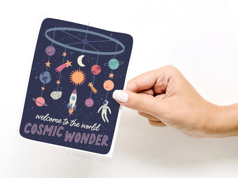A hand holds Onderkast Studio’s Welcome to the World Cosmic Wonder greeting card, featuring planets, stars, a rocket, and astronaut on a dark starry background—an ideal new baby card.