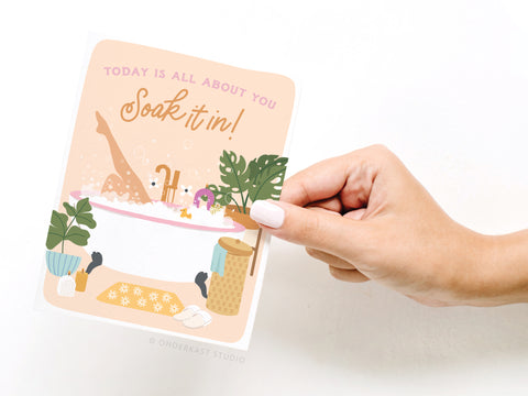 A hand holds the Soak It In Bubble Bath Greeting Card by Onderkast Studio, showing a person relaxing in a bubble bath surrounded by plants. Printed in the USA, it reads: TODAY IS ALL ABOUT YOU Soak it in! in pink and brown letters.