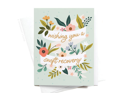 The Wishing You a Swift Recovery Greeting Card by Onderkast Studio features vibrant flowers and leaves, with two white banners displaying wishing you a swift recovery in yellow and pink script. Shown atop a white envelope.