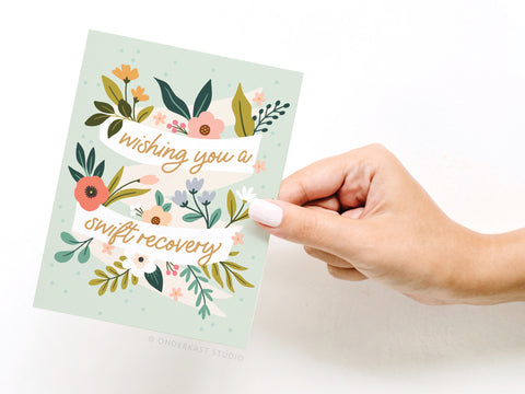 A hand holds the Onderkast Studio “Wishing You a Swift Recovery Greeting Card,” featuring colorful flowers and greenery on a light green background with the message: Wishing you a swift recovery.