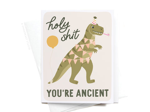 The Holy Shit You’re Ancient Dinosaur Greeting Card by Onderkast Studio features a cartoon dinosaur in party gear with the text holy shit YOURE ANCIENT—a funny milestone birthday card for anyone with a sense of humor!.