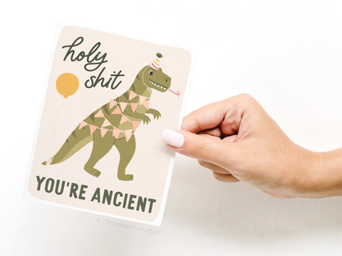 A hand holds the Onderkast Studio Holy Shit You’re Ancient Dinosaur Greeting Card, featuring a cartoon dinosaur in a party hat with decorations and balloon—a funny way to celebrate another birthday!.