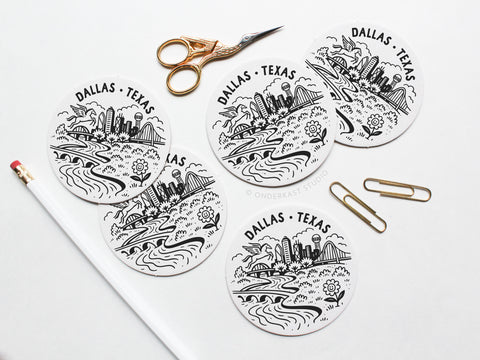 The Onderkast Studio Dallas Texas Skyline Letterpress Coaster Set includes five round coasters with a black and white cityscape design, ideal for Texas coffee tables. Displayed on a white surface with a pencil, gold scissors, and paper clips.