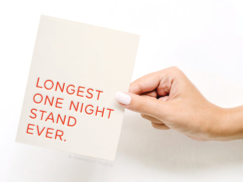 A hand with light pink nails holds the Onderkast Studio Longest One Night Stand Ever Letterpress Greeting Card, featuring bold red text and a witty anniversary message against a crisp white background.