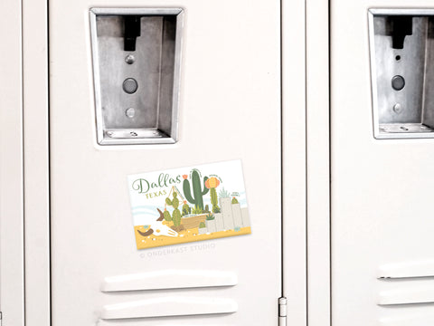 The Dallas Succulent Skyline Refrigerator Magnet by Onderkast Studio, featuring cacti, a sun, and southwestern charm, is displayed on a light-colored locker door with two other lockers partially visible in the background.