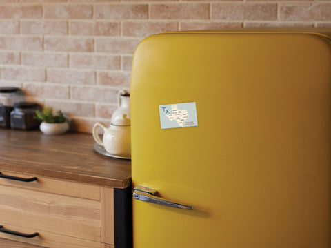 A cozy kitchen features a retro yellow fridge adorned with the Onderkast Studio Texas Map Refrigerator Magnet, wooden cabinets, a brown countertop, kitchenware, and a brick backsplash—showcasing true Texas pride.