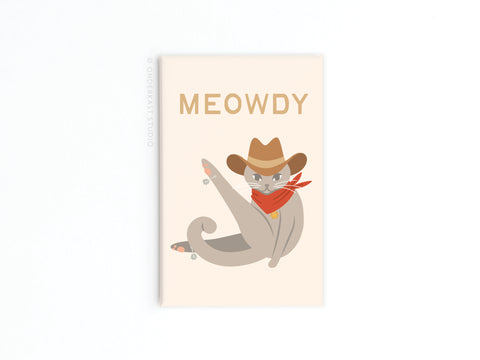 The Meowdy Cat Refrigerator Magnet by Onderkast Studio features a gray cat with Feline Flair, wearing a cowboy hat and red bandana, playfully posing under the bold “MEOWDY”—a fun addition to any fridge!.