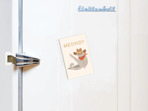 A white refrigerator with a silver handle displays the Meowdy Cat Refrigerator Magnet by Onderkast Studio, featuring a cowboy cat in a hat and bandana with the text MEOWDY.