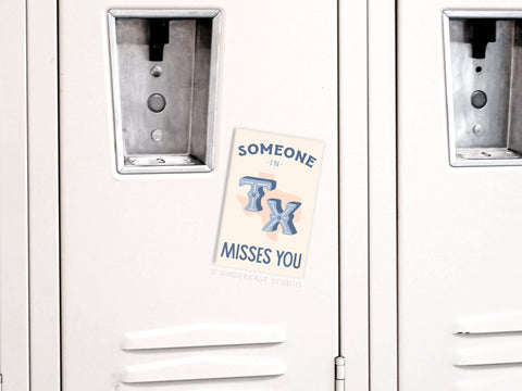 A note on a white locker reads, Someone in TX Misses You, just like the Onderkast Studio Someone in TX Misses You Refrigerator Magnet, with blue and peach letters. The closed locker doors have metal slots for locks.