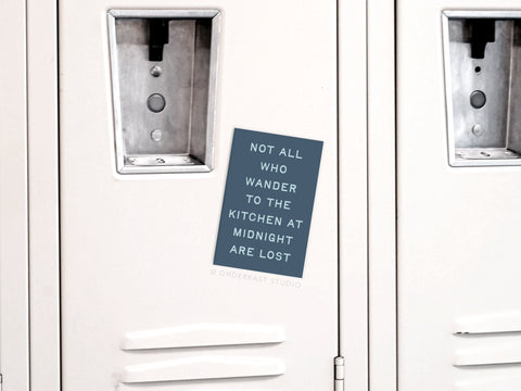 The Not All Who Wander to the Kitchen Refrigerator Magnet by Onderkast Studio holds a blue note on metallic white lockers with handles and locks. The quirky magnet reads, Not all who wander to the kitchen at midnight are lost.