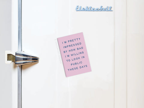 A light pink note held by the Willing to Look Bad In Public Refrigerator Magnet from Onderkast Studio is displayed on a retro white fridge. The note reads: I’M PRETTY IMPRESSED BY HOW BAD I’M WILLING TO LOOK IN PUBLIC THESE DAYS.