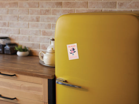 A yellow retro-style refrigerator featuring Onderkast Studio’s Bloom Where You Are Planted magnet stands in a cozy kitchen with a wooden counter, jars, a teapot set, and a brick backsplash.