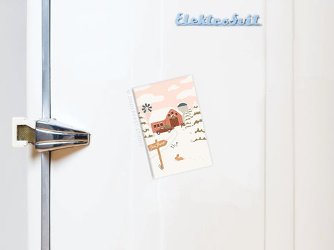 A postcard with a winter farmhouse scene adds rustic charm to a white fridge. The Christmas Tree Farm Refrigerator Magnet by Onderkast Studio holds it in place, completing the cozy display.