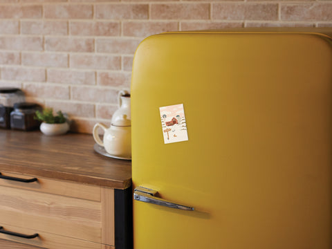 A retro yellow refrigerator with rustic charm, adorned with the Onderkast Studio Christmas Tree Farm Refrigerator Magnet, sits in a cozy kitchen with wood countertops, jars, a teapot, and a brick backsplash.