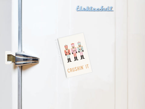 A white fridge with a metallic handle displays the Crushin’ It Nutcrackers Refrigerator Magnet by Onderkast Studio, featuring three cartoon nutcrackers and festive text—perfect for fun holiday decor and a cute touch for your kitchen.